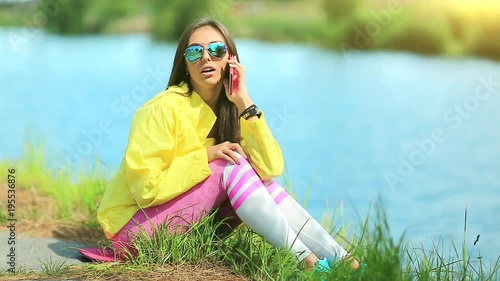 Sportswoman in yellow sports jacket sits near pond and talks on cell phone. Girl in sportswear sits near lake and speaks on smartphone photo