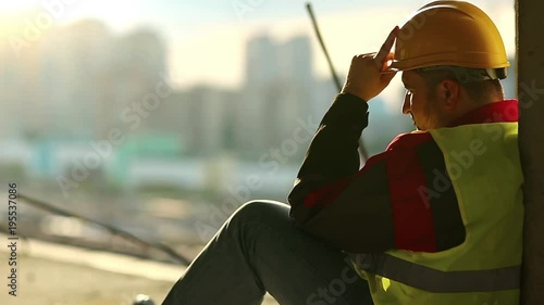Builder sits on the floor and looks ahead. Worker rests on construction site. Constructor in yellow hard hat sits on concrete floor on project site