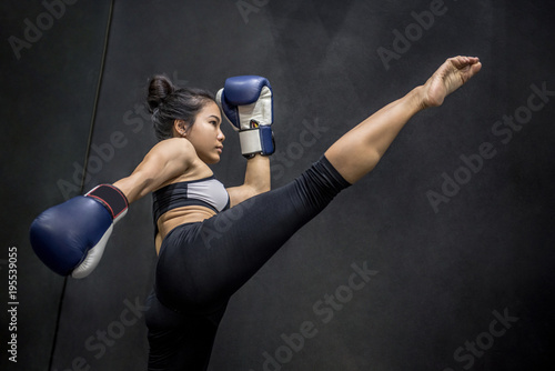 Young Asian woman boxer with blue boxing gloves kicking in the exercise gym, Martial arts on black background