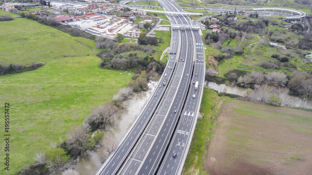 Aerial view of a stretch of highway in the Italian countryside on a cloudy day. The road is divided into three lanes in each direction. There are cars and trucks on the street. Shot in d-log.