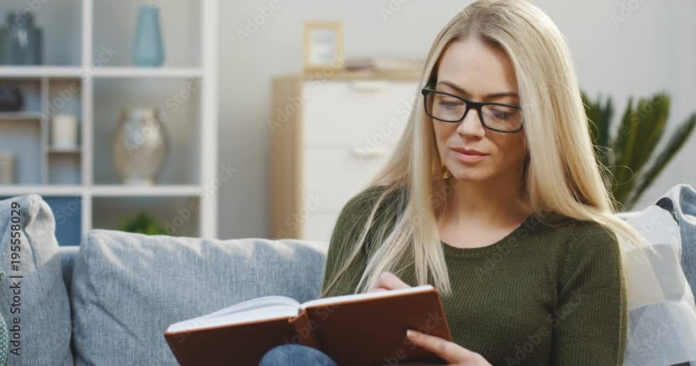 Portrait of the blonde pretty woman sitting on the sofa at home and writing notes in her notebook. Working at home. Close up. Indoor