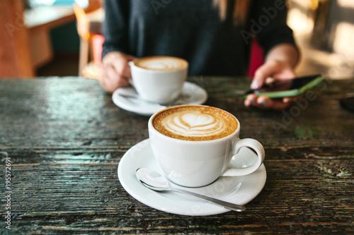 Two cups of coffee on a wooden table, a young girl holding a phone in her hand and going to call. Waiting for a meeting. Another person is late for the meeting.