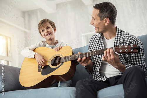 Leisure. Good-looking content little fair-haired boy smiling and holding the guitar and his father teaching him to play the guitar