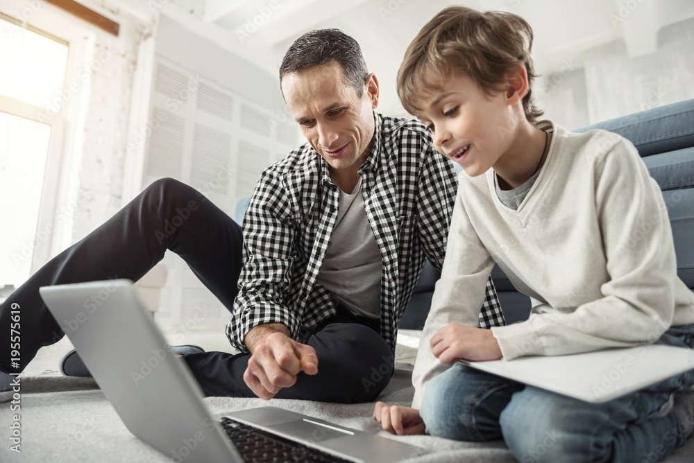 Searching the net. Good-looking inspired dark-haired man smiling and looking at the laptop and his son sitting near him on the floor Searching the net. Good-looking inspired dark-haired man smiling and looking at the laptop and his son sitting near him on the floor