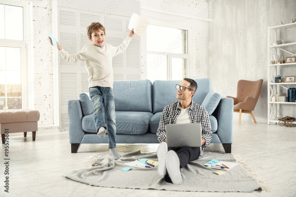 Kidding around. Attractive joyful fair-haired boy smiling and jumping while holding some papers and his father sitting on the floor Kidding around. Attractive joyful fair-haired boy smiling and jumping while holding some papers and his father sitting on the floor