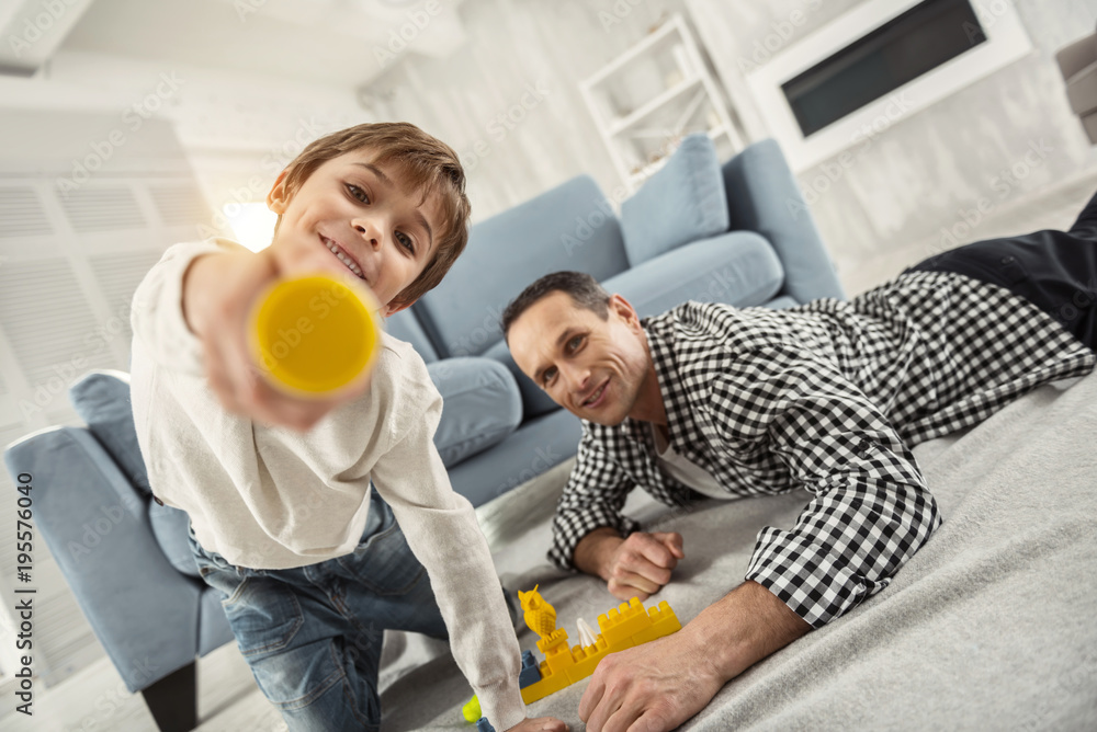 Happy childhood. Attractive inspired fair-haired boy playing with his erector set and his daddy lying near him and playing with him Happy childhood. Attractive inspired fair-haired boy playing with his erector set and his daddy lying near him and playing with him
