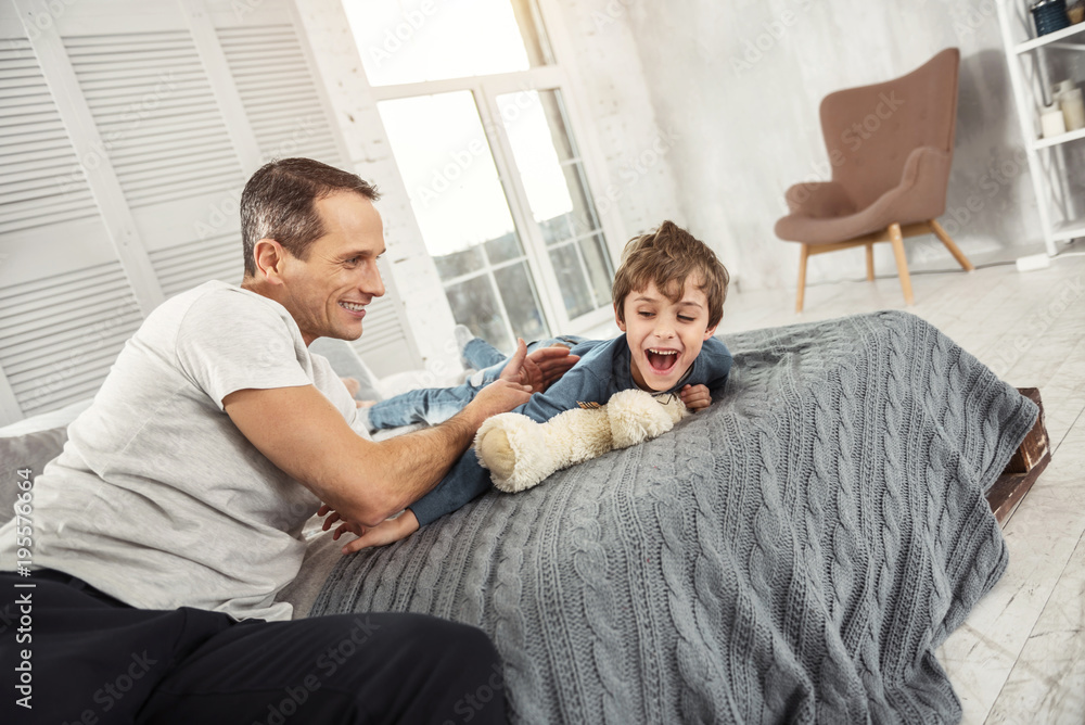 Having fun. Nice exuberant fair-haired boy laughing and lying on the bed and his daddy playing with him Having fun. Nice exuberant fair-haired boy laughing and lying on the bed and his daddy playing with him