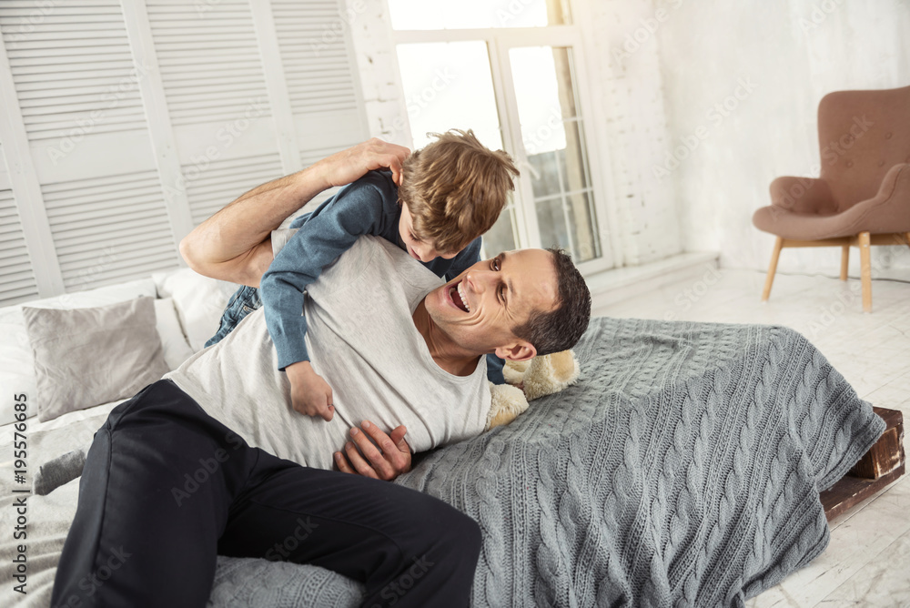 Amazing day. Handsome alert dark-haired daddy laughing and lying on the bed with his son and they having fun Amazing day. Handsome alert dark-haired daddy laughing and lying on the bed with his son and they having fun