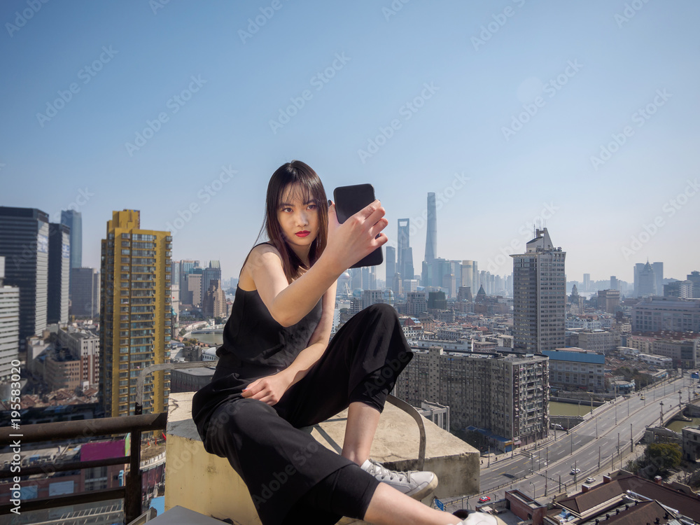 Fototapeta premium Beautiful young brunette woman in black dress sit on top of mansion roof and take selfie photo with blur Shanghai Bund landmark buildings background. Emotions, people, beauty and lifestyle concept.
