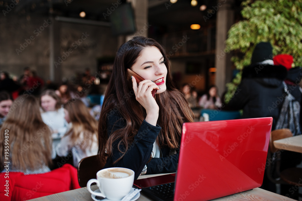Brunette girl sitting on cafe, working with red laptop and speaking at mobile phone. Brunette girl sitting on cafe, working with red laptop and speaking at mobile phone.