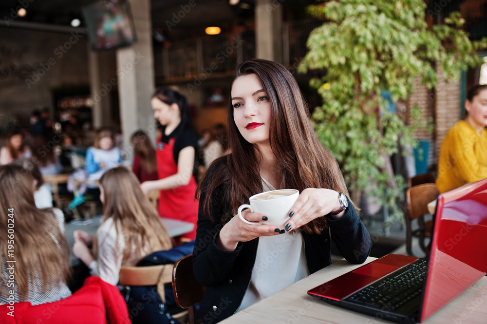 Brunette girl sitting on cafe with cup of cappuccino, working with red laptop.