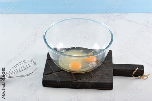 In a deep bowl, eggs, sugar, vanillin for biscuit. In the frame, a whisk for knocking. Light marble background. Close-up. photo