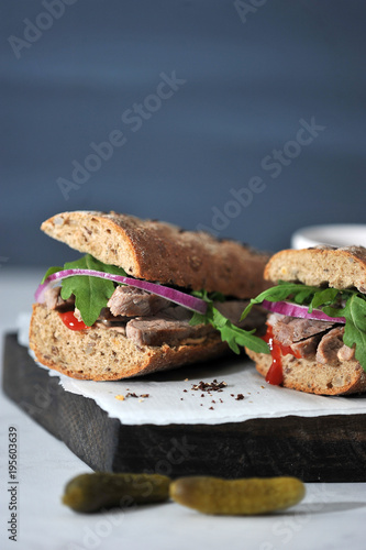 Sandwich on a wooden board. The filling of the sandwich consists of beef, arugula, red onion and marinated cucumber slices. Near the sandwich two cucumbers. Vertical shot. Negative space.