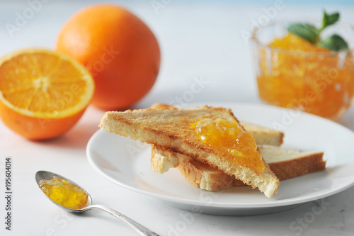Orange jam and plate with toast. In the frame, an orange and half an orange. One toast is smeared with jam. Next to the plate is a teaspoon with jam. Light background. Close-up. Macro photography.