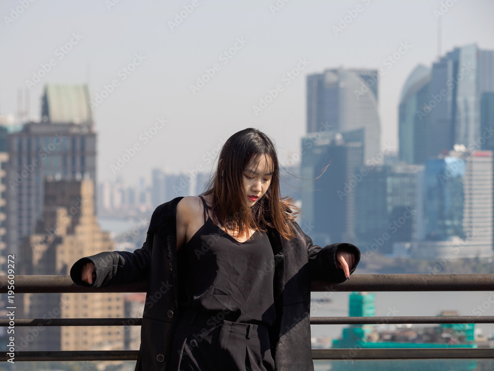 Beautiful young brunette woman in black jacket stand on top of mansion roof and lower her head in wind with Shanghai Bund landmark buildings background. Emotions, people, beauty and lifestyle concept.