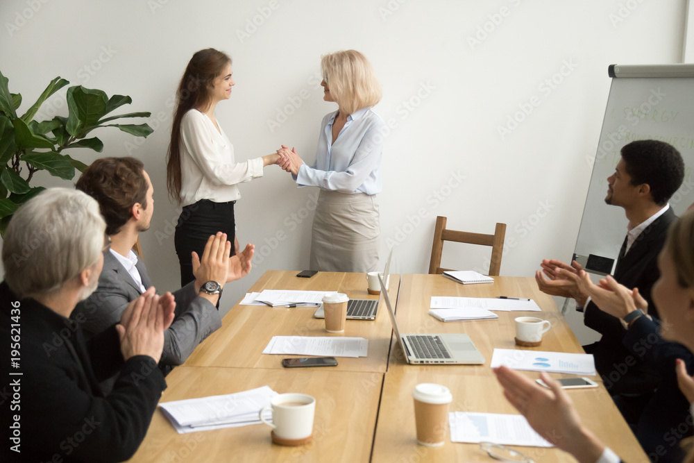 Senior businesswoman boss promoting female employee thanking for good work holding hands as trust and support concept while team applauding congratulating colleague at group meeting, recognition