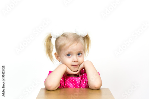 A beautiful young blond girl leans her elbows on a chair and puts her head on her hands. Little girl 3 years old on a white background put her head on her hands.