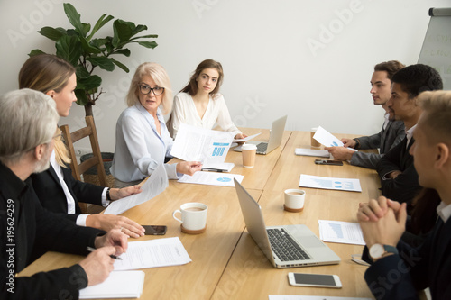 Serious aged businesswoman discussing corporate financial report with team talking to employees, senior mature female boss ceo leader analyzing work results with subordinates at company group meeting