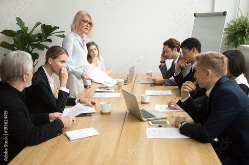 Dissatisfied senior woman boss scolding employees for bad work at diverse group meeting, angry female executive team leader reprimanding subordinates for poor financial result at office briefing