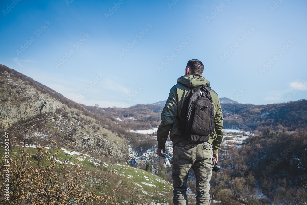 Young man standing on top of cliff in winter mountains holding a camera and enjoying view.