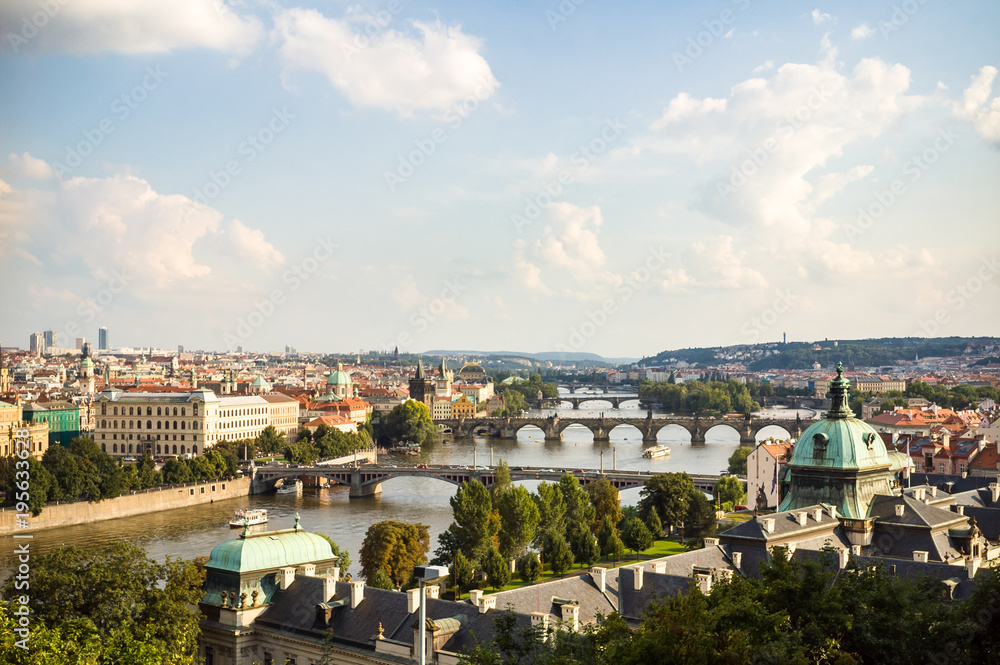 Panoramic view of the Charles bridge on the river Vltava in the historic center of Prague from the Letna park with copper rooftops in the foreground under a pale blue sky at the end of the day.