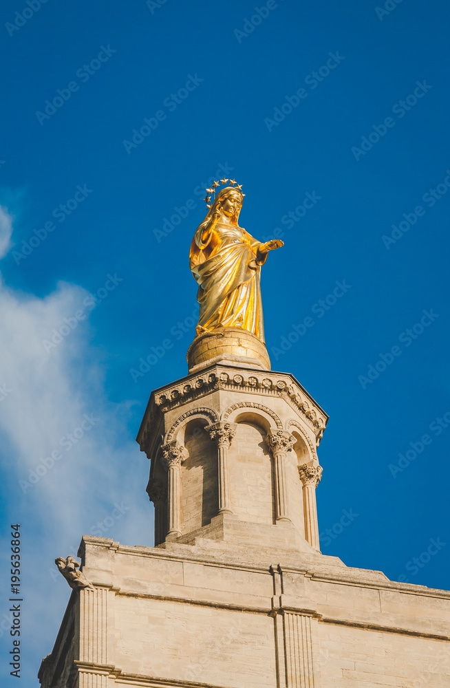 Golden statue of Blessed Virgin Mary against blue sky. It is atop bell tower of Cathedral of Notre-Dame des Doms built in 12th century in Avignon, France