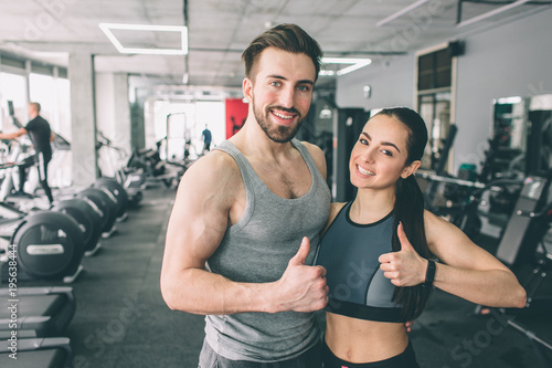 Trainer and her student are standing in the sport club's training room together and showing their big thumb's up. They are happy to exsercise in this fitness club. Young couple is smiling.