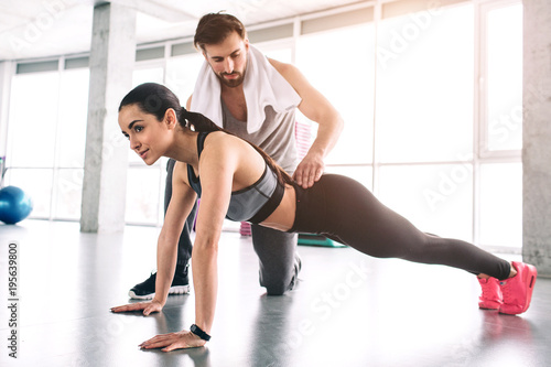 Trainer and his student are in a fitness room. She is doing high plank exercise while he is controlling the quality of what she is doing.