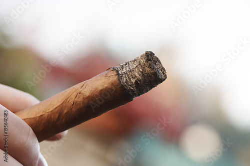 A hand holding a burning Cuban cigar with a bokeh
