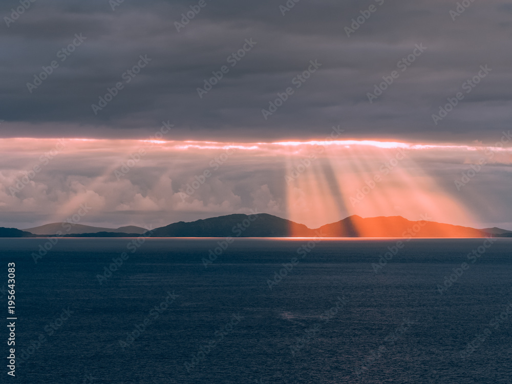 Sunset over the Outer Hebrides, as seen from the Neist Point cliff on the Isle of Skye, Scotland. Dramatic rays of light beaming through the dark clouds and shine in the deep blue sea on a summer day. Sunset over the Outer Hebrides, as seen from the Neist Point cliff on the Isle of Skye, Scotland. Dramatic rays of light beaming through the dark clouds and shine in the deep blue sea on a summer day.