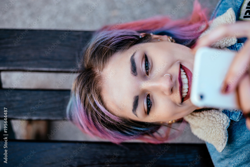 Close-up portrait of a smiling girl with dyed hair using phone while lying on a bench. Top view. - obrazy, fototapety, plakaty 