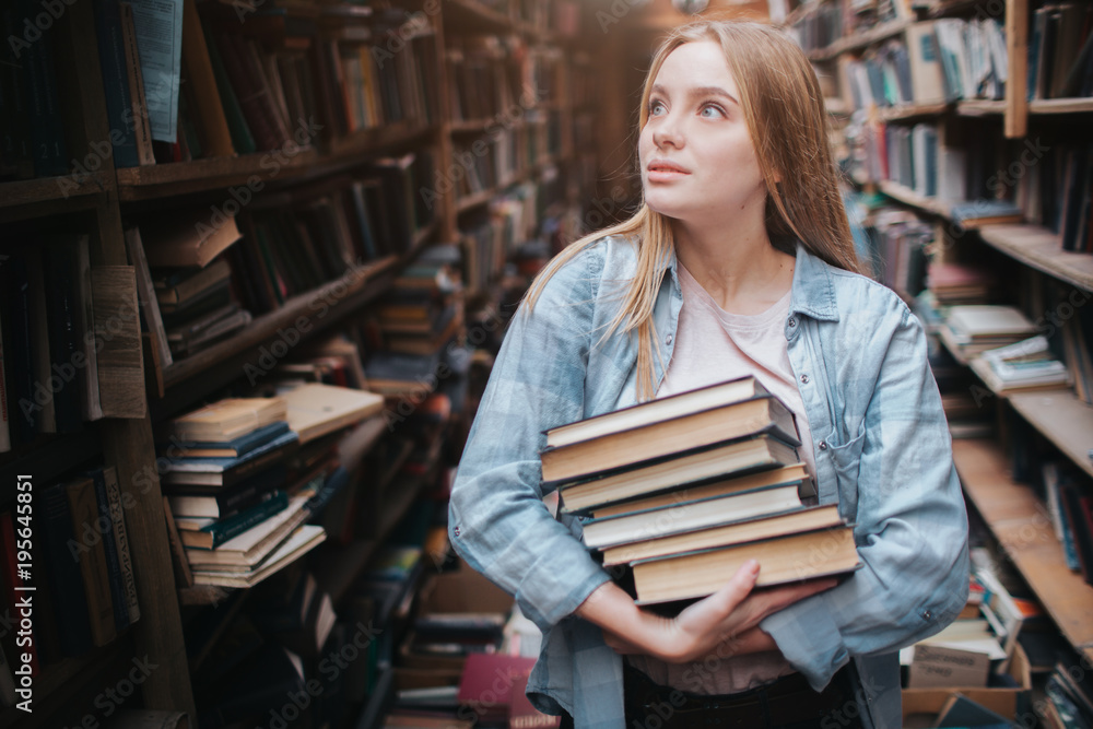 A small girl standing in a big old book store and holding many books in her hands. She is searching for another book to take with her. Young beauty looks dreamy. A small girl standing in a big old book store and holding many books in her hands. She is searching for another book to take with her. Young beauty looks dreamy.