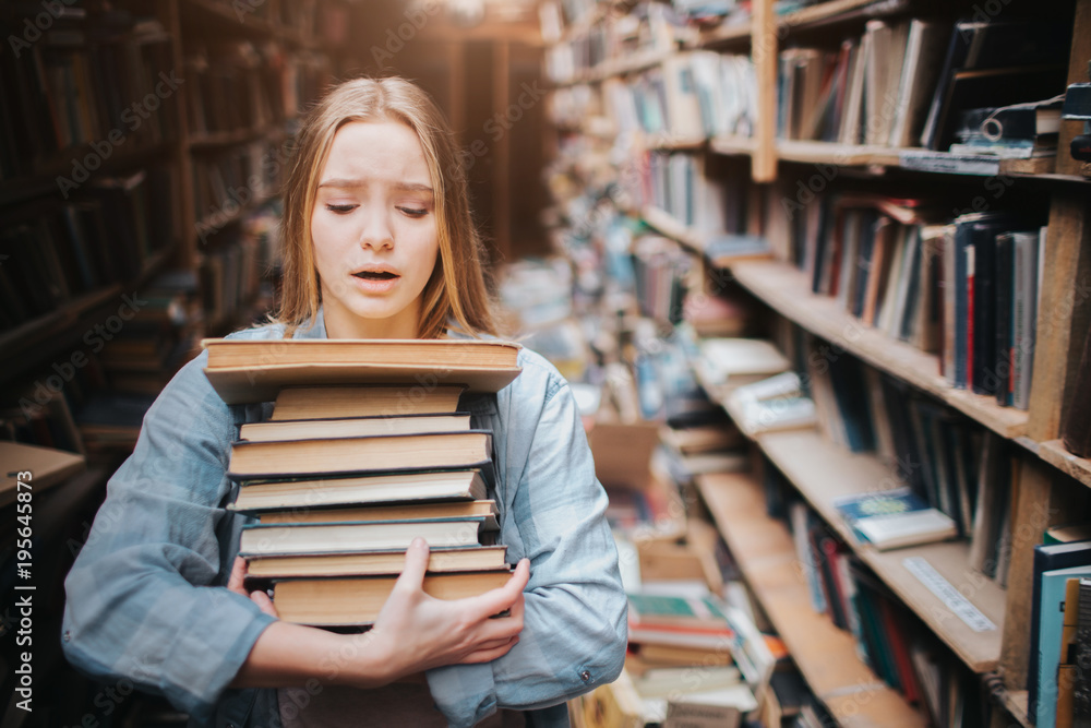 Girl is carrying a lot of books in her hands. It is hard for her to do. She looks hepless and tired. The girl stands in big old library. Girl is carrying a lot of books in her hands. It is hard for her to do. She looks hepless and tired. The girl stands in big old library.