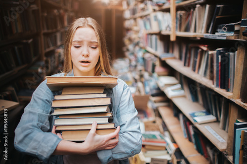 Girl is carrying a lot of books in her hands. It is hard for her to do. She looks hepless and tired. The girl stands in big old library.