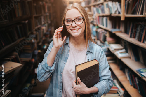 A picture of attractive girl standing with a book in her left hand and talking on the phone. She is looking straight forward and smiling. This woman is very nice.