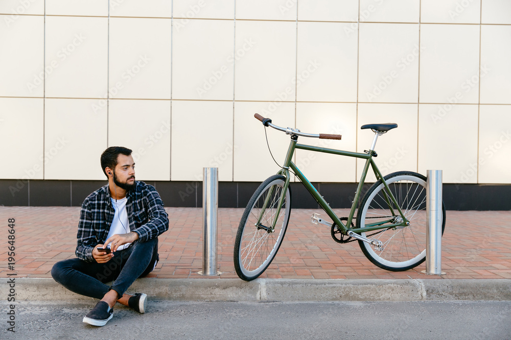 Obraz premium Young bearded man listening to music in earphones on his smartphone while sitting on the ground near the bike, outdoors. Wearing stylish clothes.