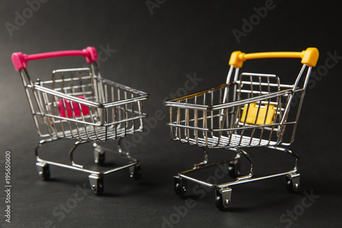 Close up of two supermarket grocery push carts for shopping with wheels and yellow and pink plastic elements on handle isolated on black background. Concept of shopping. Copy space for advertisement