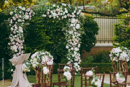 Beautiful decoration of a wedding ceremony in a green autumn garden. Brown wooden chairs for guests on either side of the walkway, which leads to a festive arch decorated with flowers