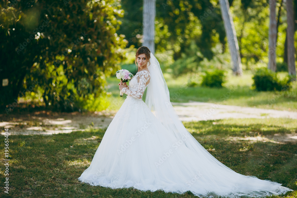 Beautiful wedding photosession. The bride in a white lace dress with a long plume, hairpin in hair, veil and bouquet of white and pink flowers in a large green garden on weathery sunny day
