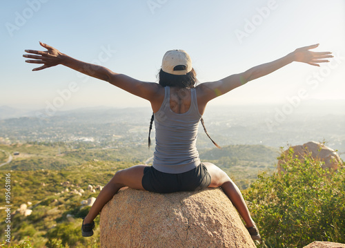 fit female hiker sitting on rock at mountain top looking at city in distance with arms up happy with accomplishment at sunset