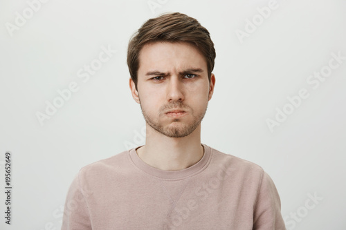 You disappointed me. Indoor portrait of handsome unshaven young man sulking and frowning eyebrows, being offended with cruel words, standing against gray background. Son waits for apology