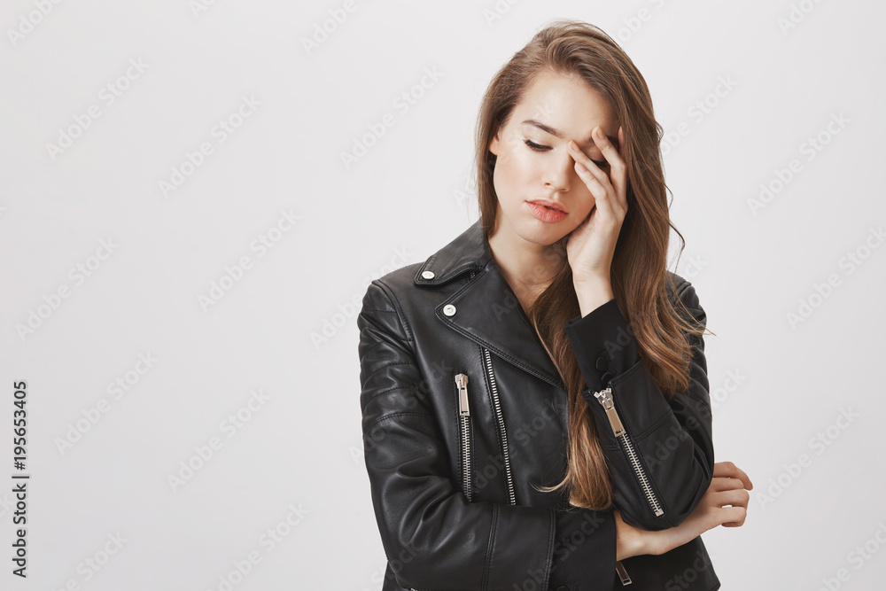 Studio shot of gloomy feminine urban woman in leather jacket looking down and touching forehead with hands, feeling miserable or exhausted, having headache from stress, standing over gray background Studio shot of gloomy feminine urban woman in leather jacket looking down and touching forehead with hands, feeling miserable or exhausted, having headache from stress, standing over gray background