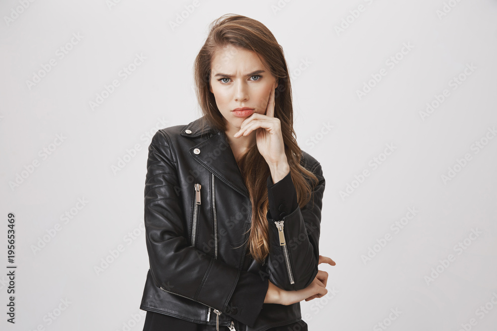 Offended gloomy girlfriend waiting for apology. indoor shot of good-looking urban woman in trendy leather jacket, frowning, holding hand on face, staring with contempt at camera over gray background Offended gloomy girlfriend waiting for apology. indoor shot of good-looking urban woman in trendy leather jacket, frowning, holding hand on face, staring with contempt at camera over gray background