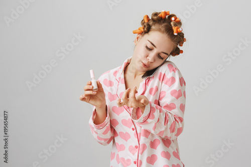 Treat yourself like queen. Portrait of attractive young woman in hair curlers and pyjamas, holding smartphone on shoulder while talking with friend, painting nails, and blowing on it to dry faster