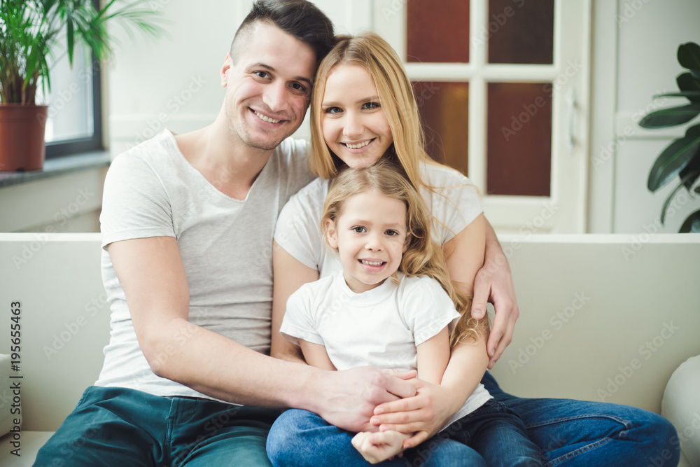 Happy young beautiful smiling parents with a little blond daughter embrace and play sitting at home on the couch. The concept of family Happy young beautiful smiling parents with a little blond daughter embrace and play sitting at home on the couch. The concept of family