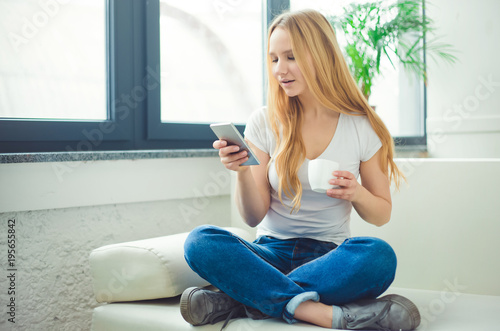 Portrait of a beautiful young blond woman sitting at home on the couch with a phone. Relax at home. Communication with friends on the Internet