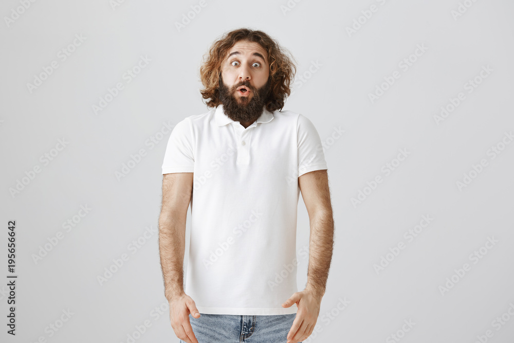 Studio shot of handsome easter man with curly hair and beard standing shocked with dropped jaw and popped eyes, being astonished with kid that jumped from corner and scared him over gray background Studio shot of handsome easter man with curly hair and beard standing shocked with dropped jaw and popped eyes, being astonished with kid that jumped from corner and scared him over gray background