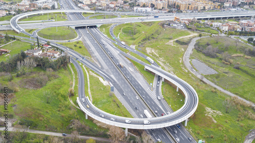 Aerial view of a series of ramps of the Rome motorway junction. Many cars, motorcycles and trucks drive on the road. Around there are grass, meadows and green trees.