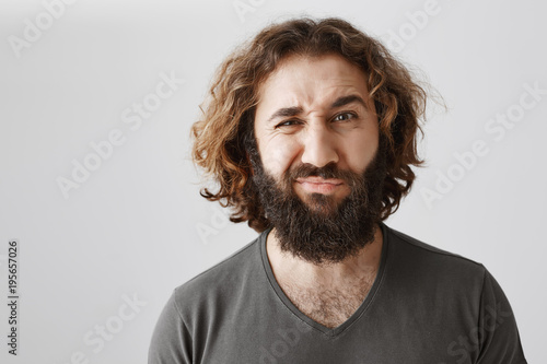 Man is not sure, having doubt. Portrait of attractive curly-haired husband frowning and grimacing from dislike and displeasure, hearing something ridiculous or dumb while standing over gray wall