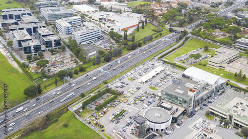 Aerial view of a stretch of highway in the Italian countryside. The road is divided into many lanes in each direction. There are cars and trucks on the street.Around the road there is industrial area.